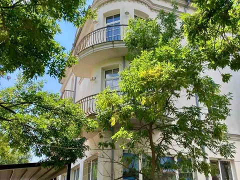 Ъгловата фасада на сградата с балкони, полускрита зад зелени дървета