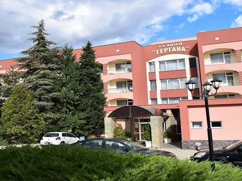 Розовата фасада на хотел „Гергана“ с вход, борове и паркирани коли