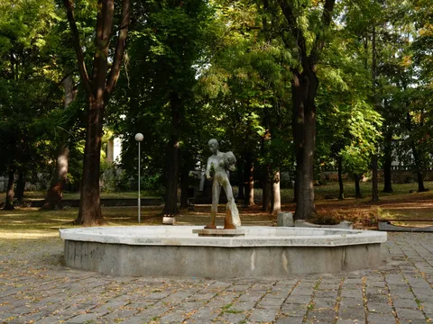 Статуята на голо момче в празния басейн на фонтана, около нея високи дървета
