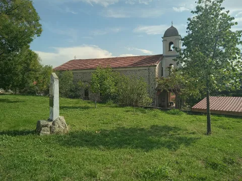 Храмът „Успение Богородично“ и камбанарията сред тревистия двор с паметна колона