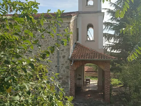 Камбанарията и входът на храма „Успение Богородично“ сред зеленина