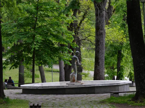 Каменна алея към кръглия фонтан със статуя сред високите дървета в парка