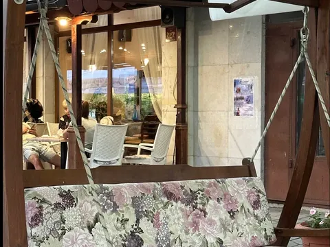 Дървена люлка с флорална възглавница пред остъкления салон на „Старата пицария“