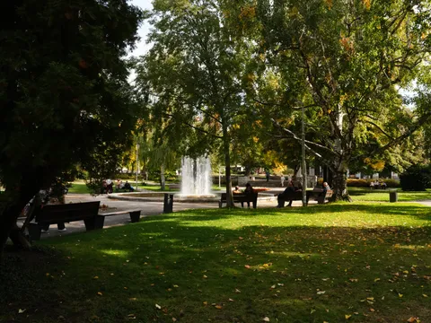 Поляна под дърветата, в дъното фонтанът и хора на пейки