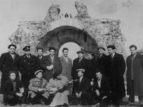 Стара снимка: група мъже в палта, единият със струнен инструмент, пред Камилите