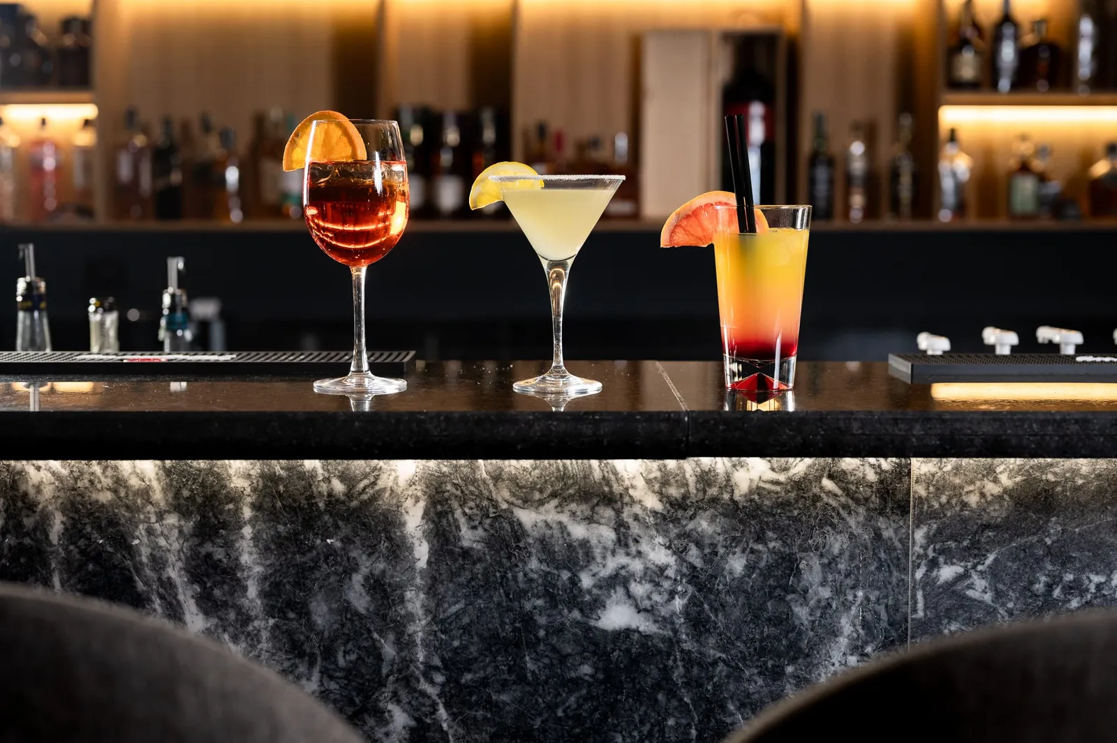 Три коктейла на мраморния бар на Senate Bar & Dinner пред осветени рафтове с бутилки