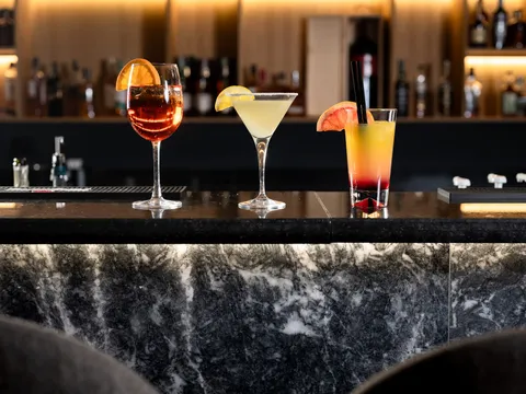 Три коктейла на мраморния бар на Senate Bar & Dinner пред осветени рафтове с бутилки