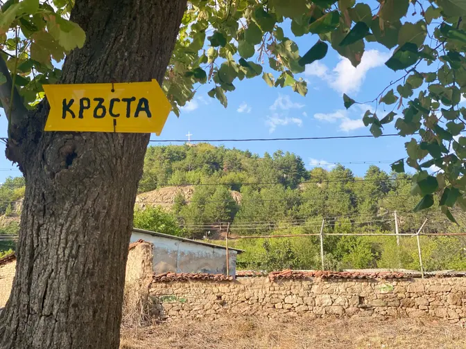 Жълта табела „Кръста“ на дърво, а на хълма отсреща се вижда кръстът